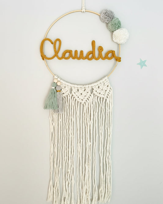 atrapasueños con el nombre de Claudia en tono mostaza, tres pompones gris, verde y blanco y dos borlas verde y gris abajo a la izquierda. decorado con flecos con dibujo macramé